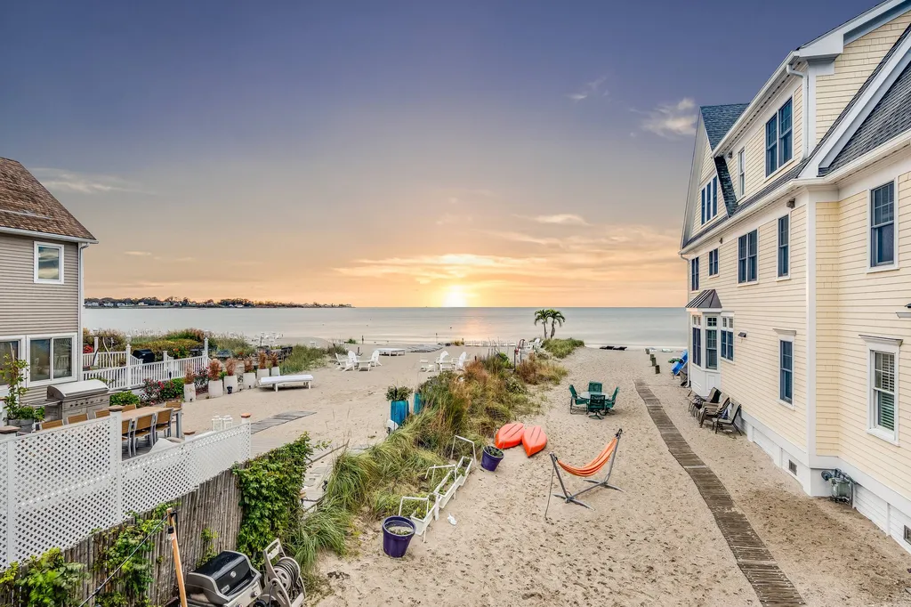 Sandy waterfront between neighboring houses, with kayaks, an orange hammock and the sun low over the water.